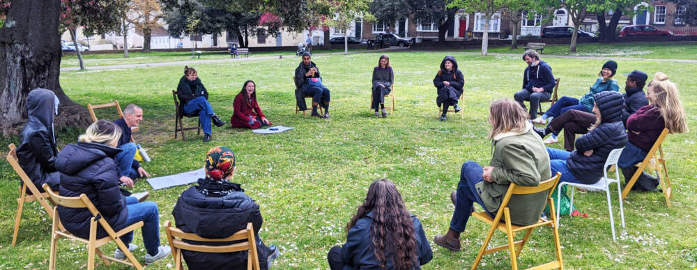 Art school students sit on chairs in a large circle on a green grass public space under trees