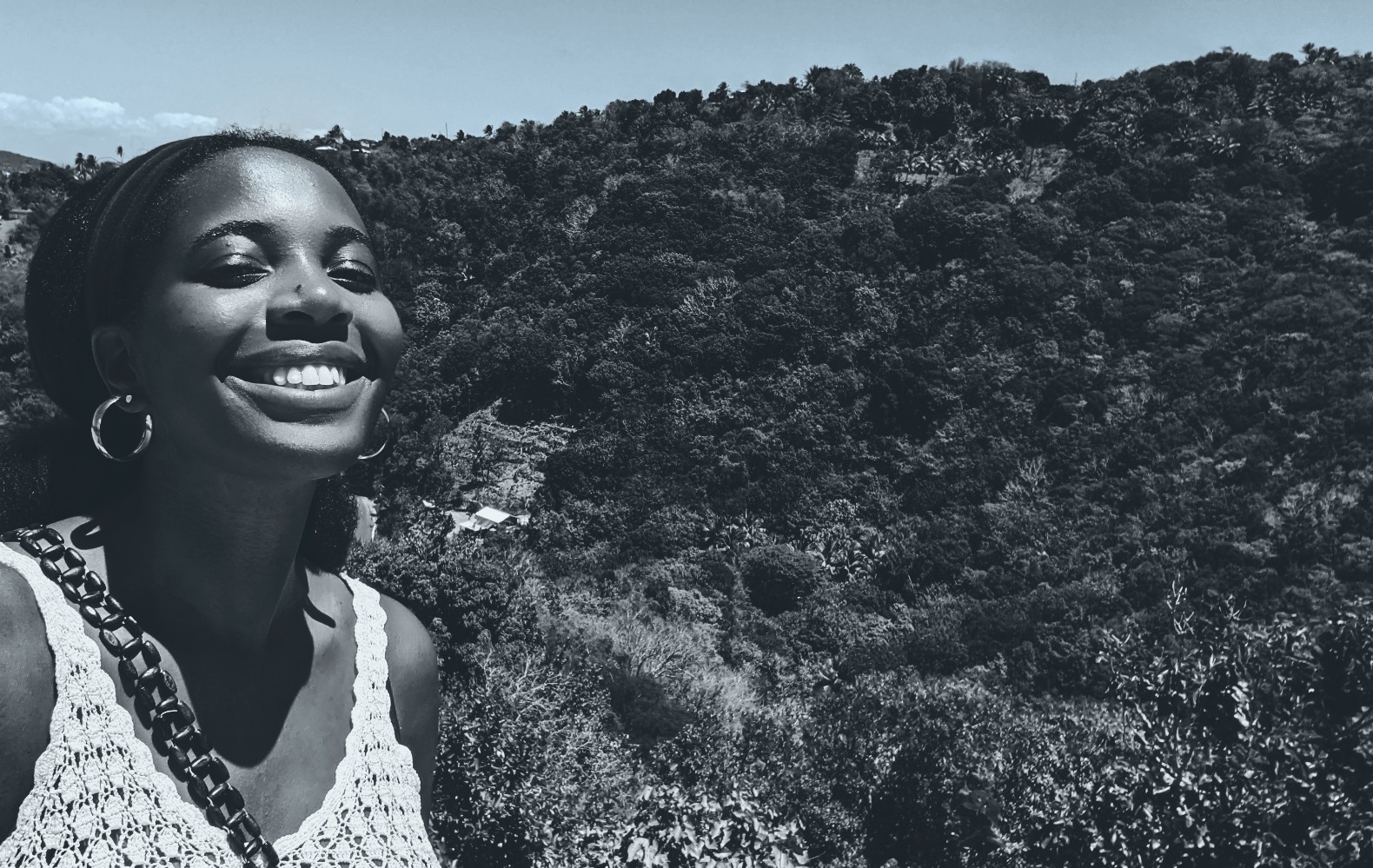 Jaccaidi smiles against a mountain view with lush vegetation, the image is black and white but is sunny