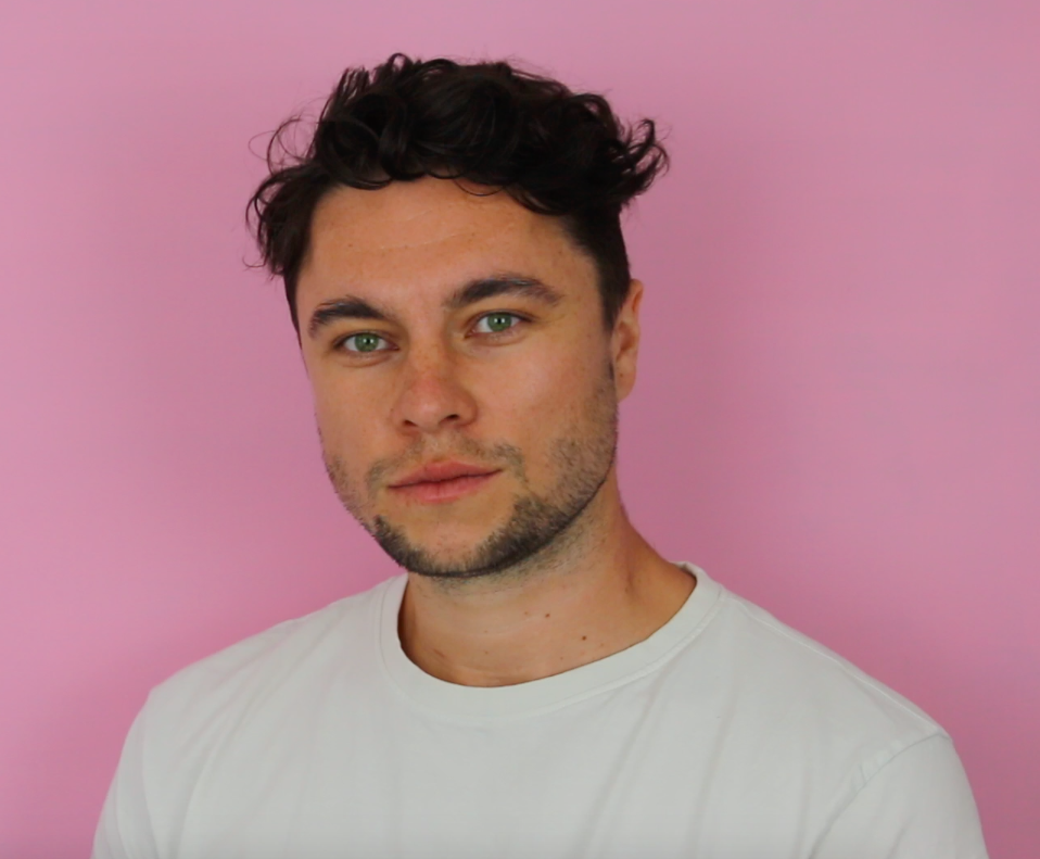 A head and shoulders shot of Kieran who has brown hair, wearing a white tee shirt and infront of a pink background.