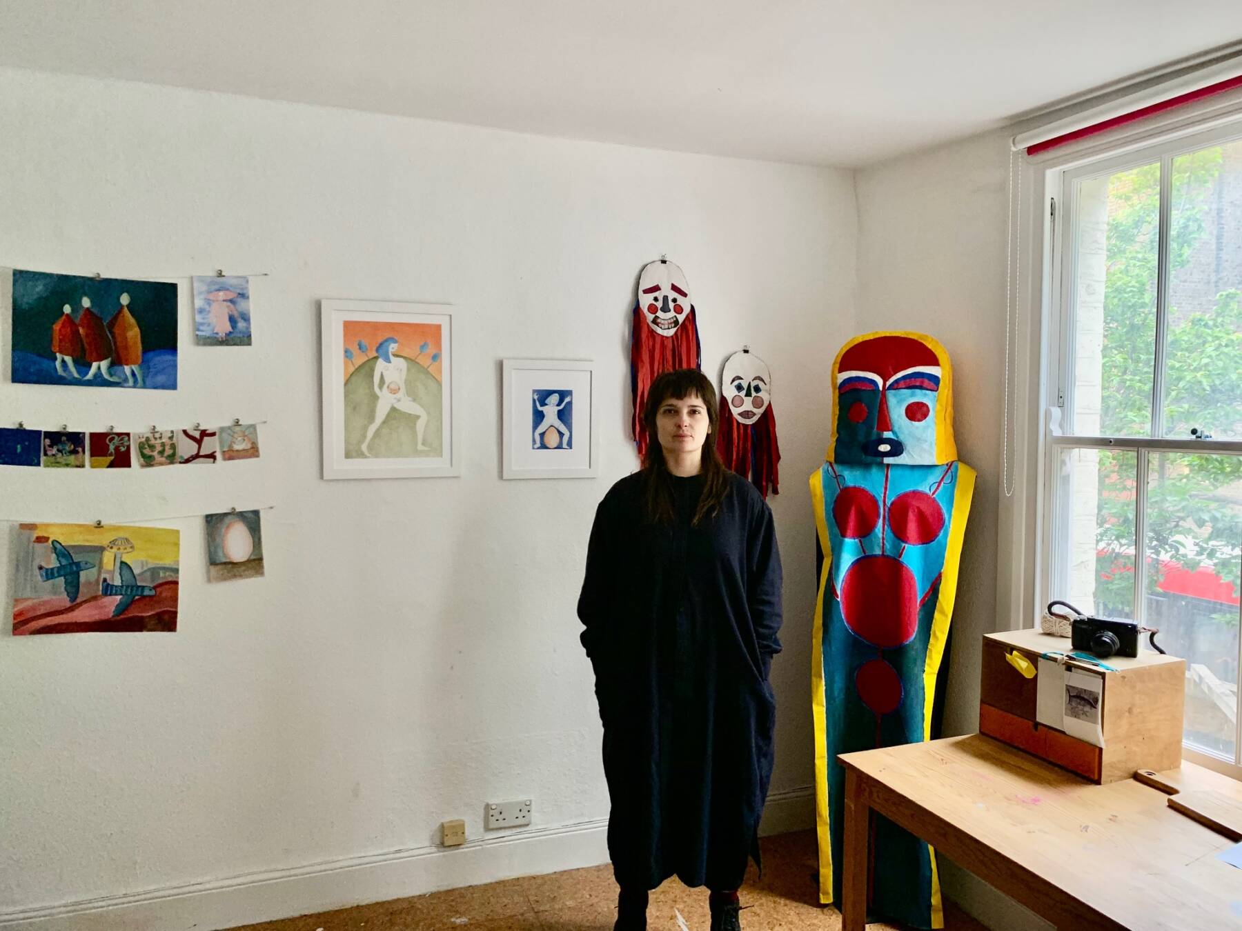 Milou Stella stands in a room with colorful artowrk and a costume in the corner of the room. They wear balck and have black hair with a fringe.